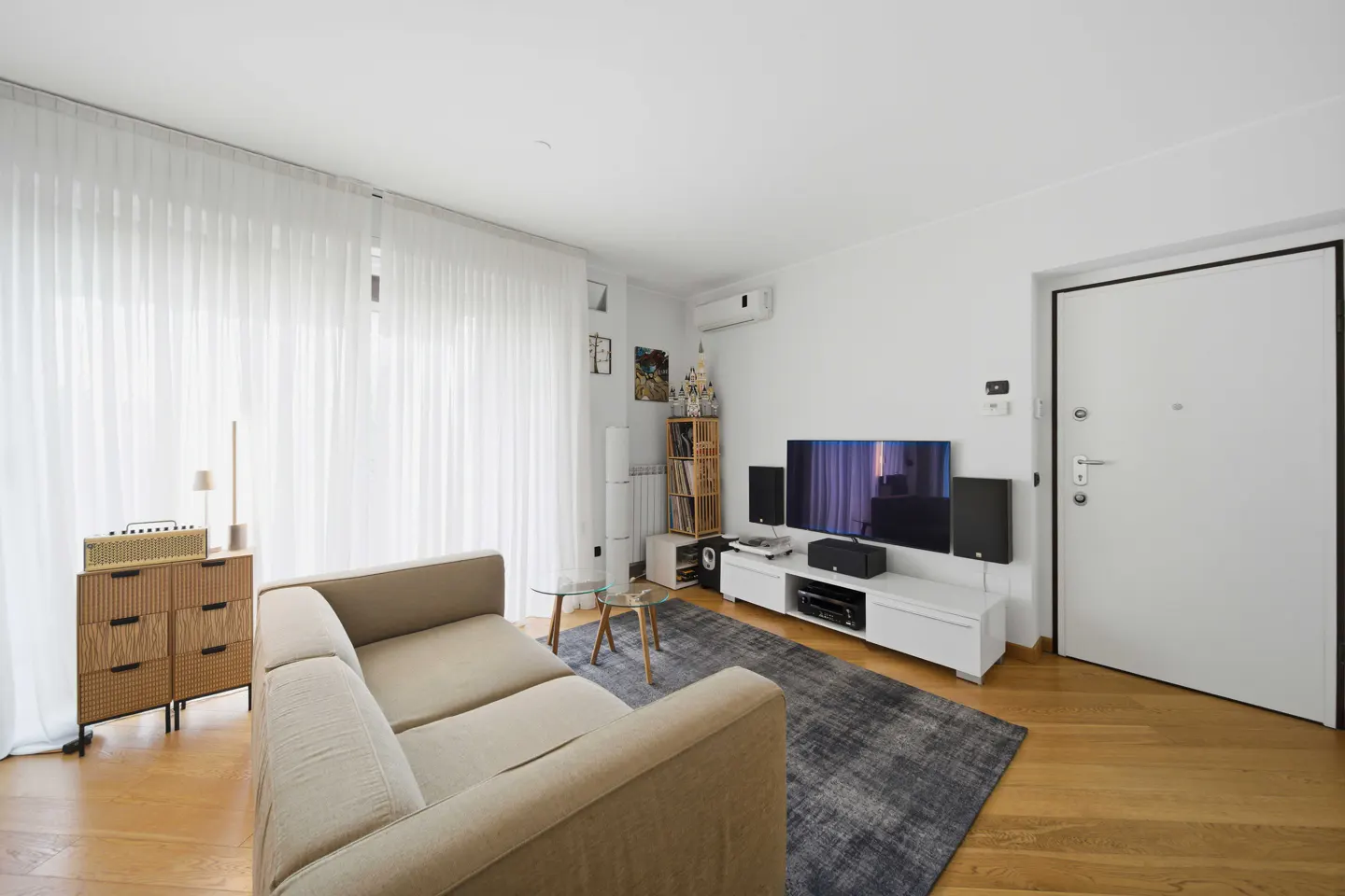 Living room with beige sofa, white walls, and light wood floors. A TV sits on a white console with speakers. Sheer white curtains cover a large window.