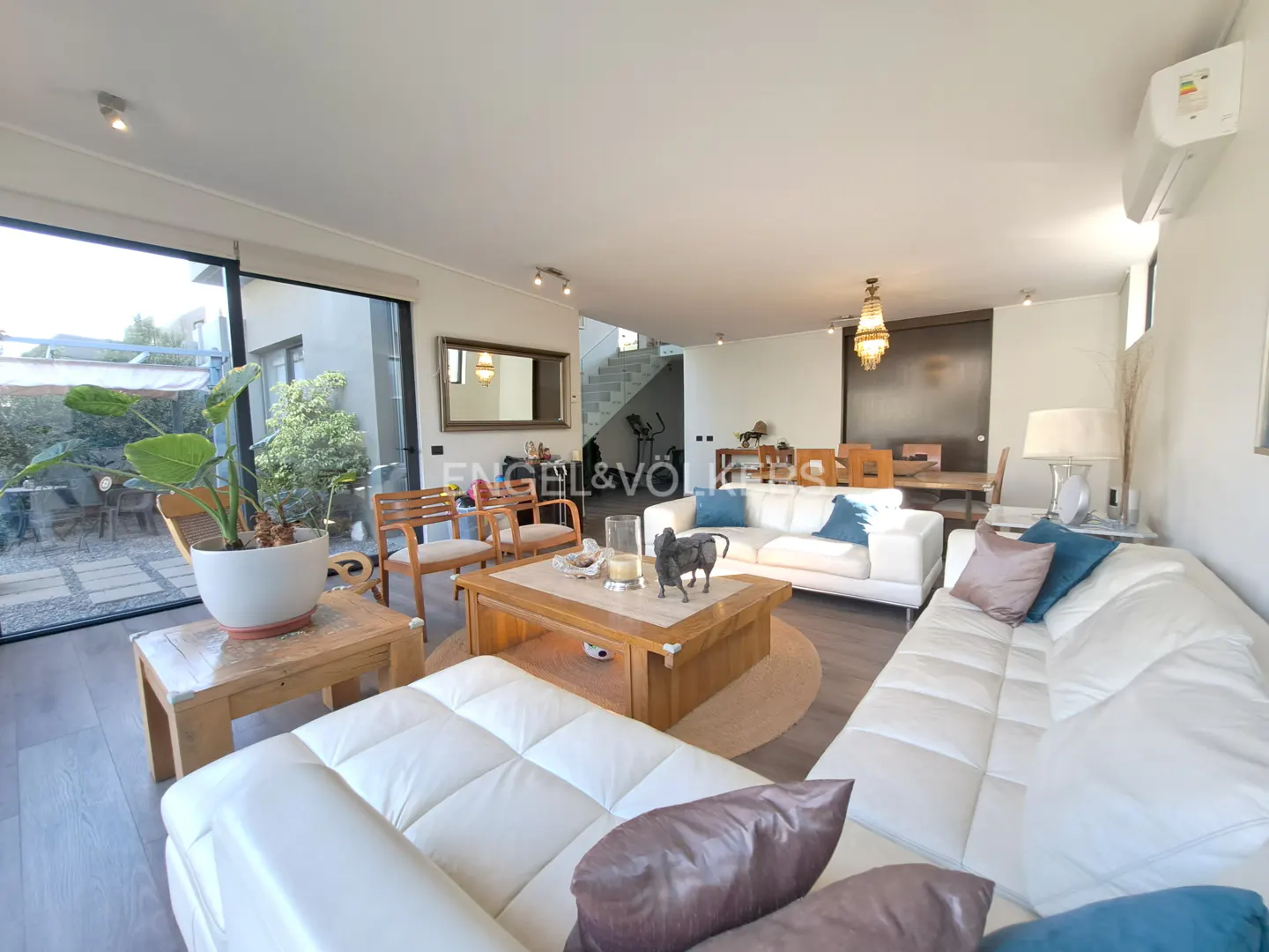 Bright living room with white sofas, wood furniture, and a view of a garden through a large window. Engel & Völkers logo visible.