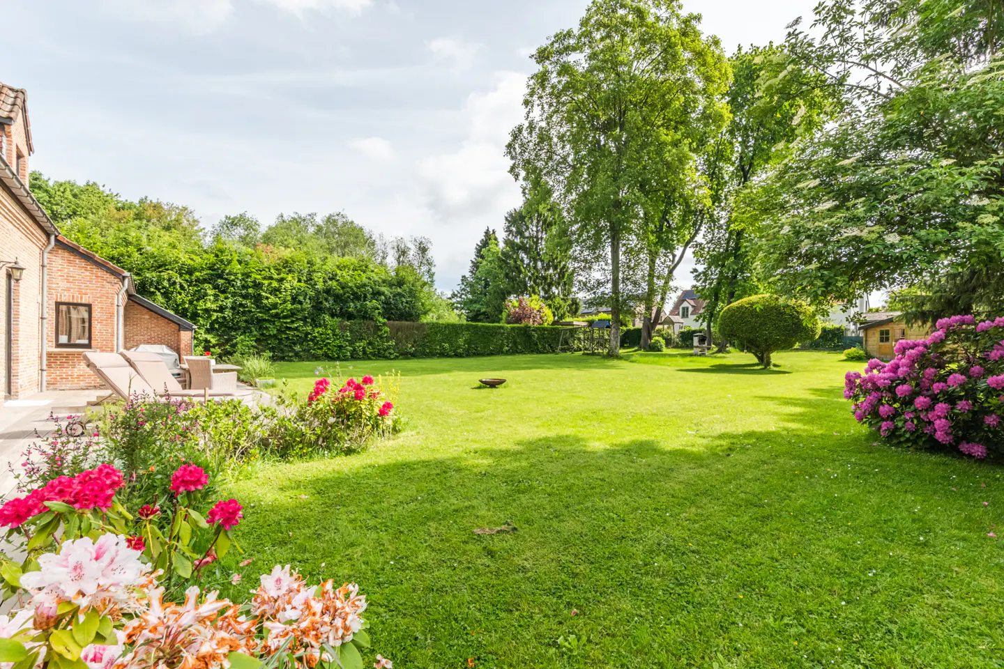 A large green lawn with trees and flowers, next to a brick house with patio furniture.