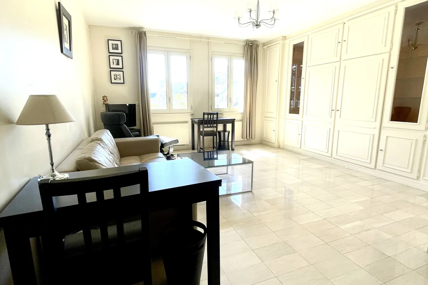Bright living room with beige walls, tile floor, and built-in white cabinets. A tan sofa, black tables, and dining set furnish the space.