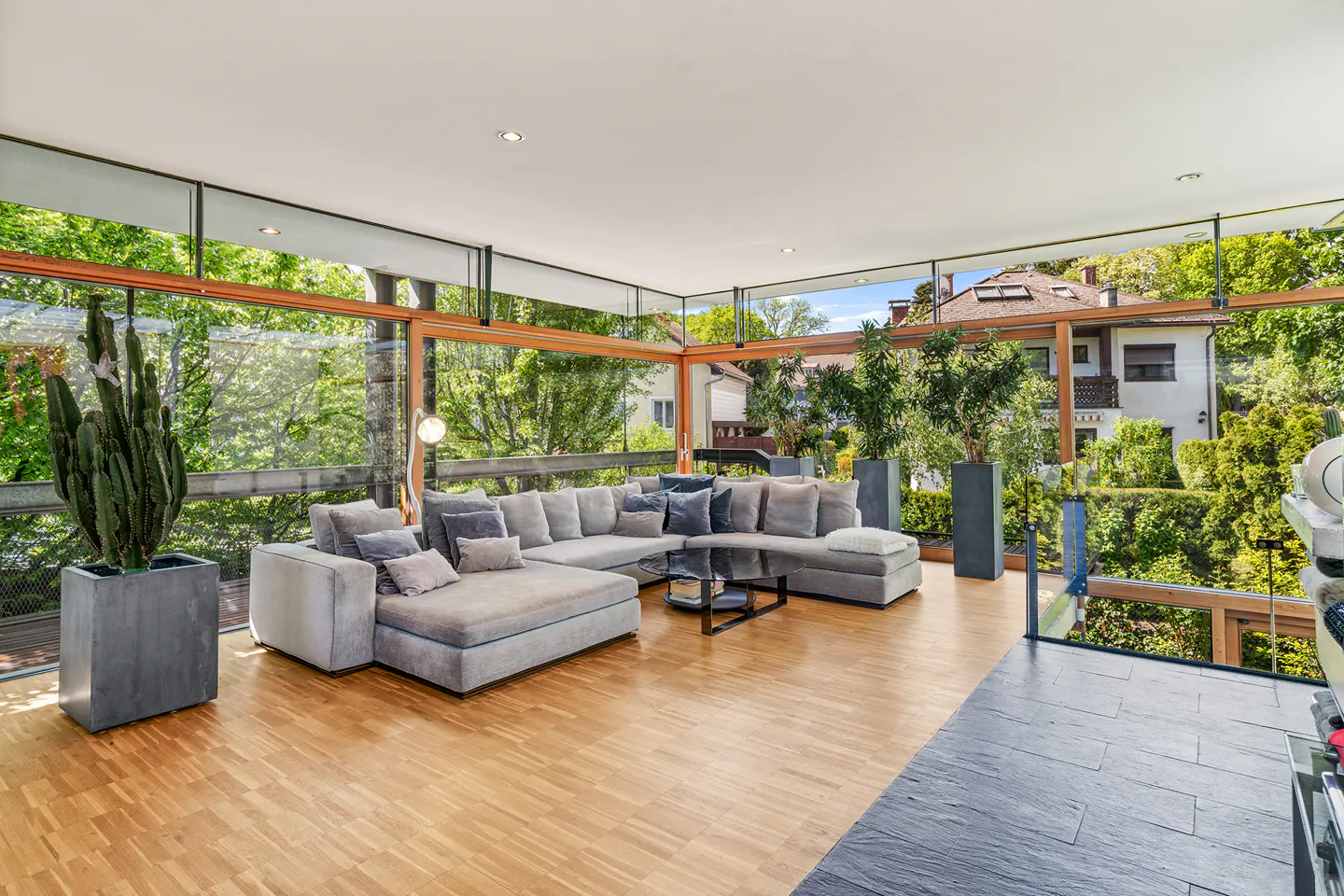 Bright living room with wood floors, a gray sectional sofa, and floor-to-ceiling windows overlooking lush greenery.
