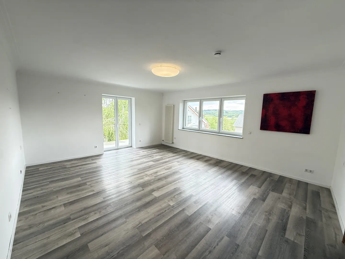 Bright, empty room with gray wood-look floors, white walls, and a modern light fixture. Windows and glass doors offer outdoor views. A red abstract painting hangs on the wall.