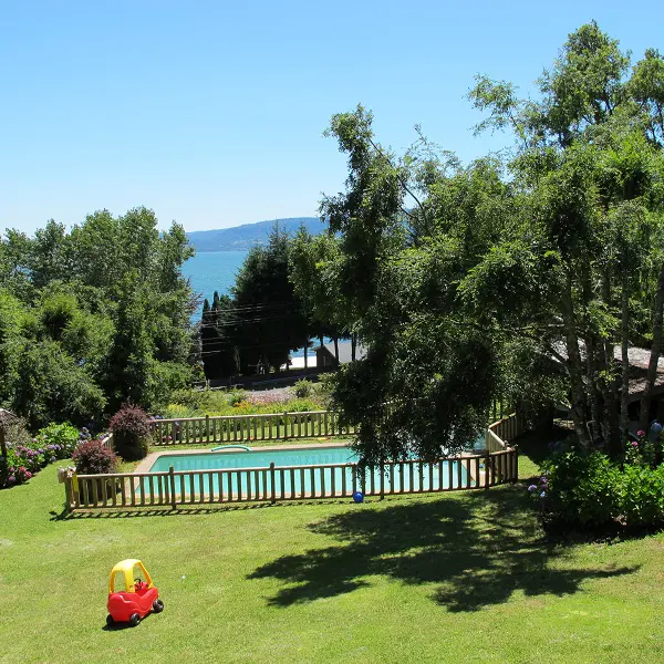 A backyard with a pool, trees, and a red and yellow toy car on the green grass. In the background is a blue lake and sky.