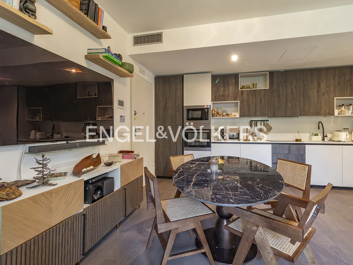 A modern apartment interior with a black marble table and wooden chairs. The kitchen has white and wood cabinets. A large TV is on the left.