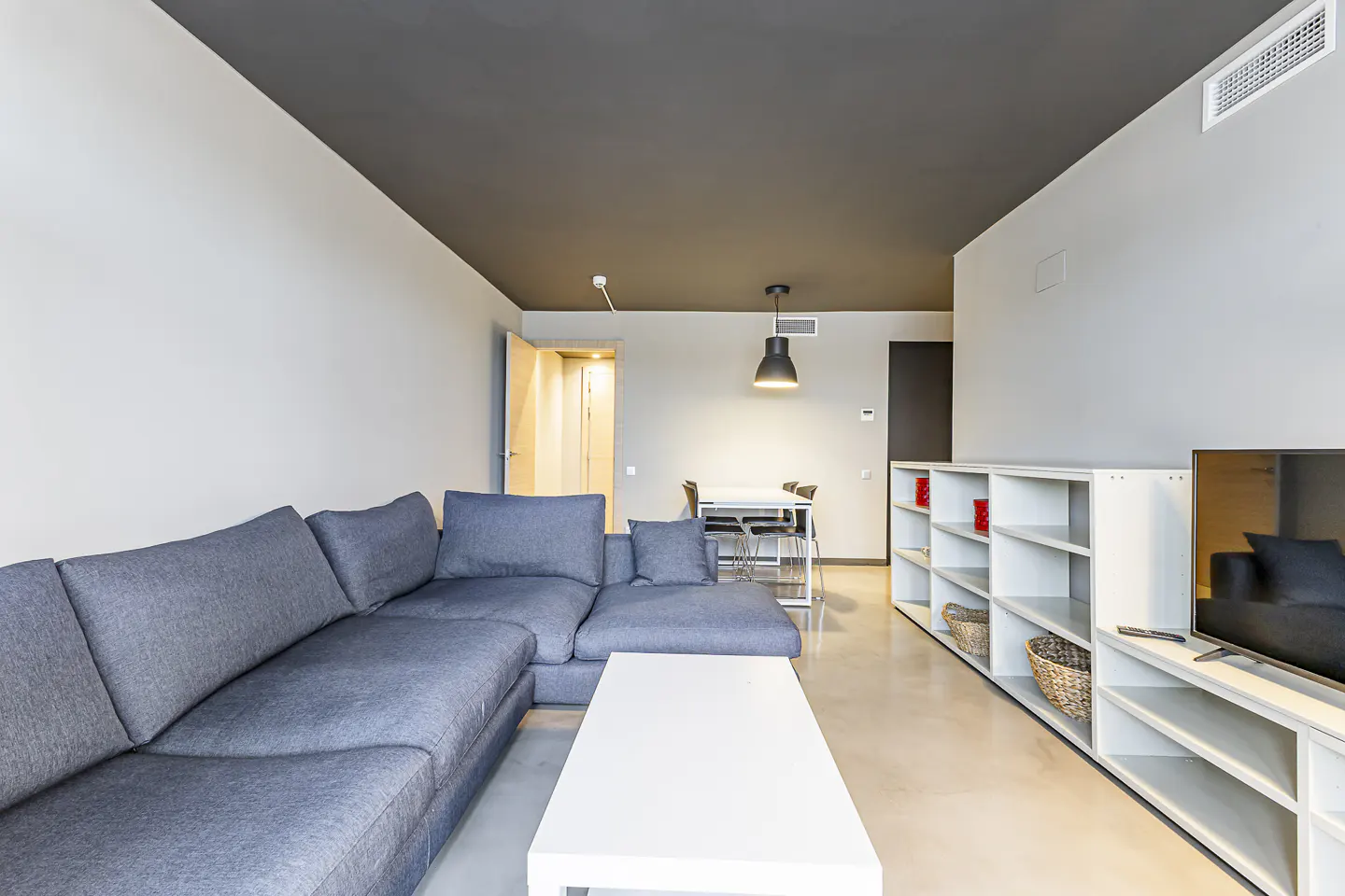 Living room with gray sectional sofa, white coffee table, and white shelving unit with TV. Dining table and chairs are in the background.