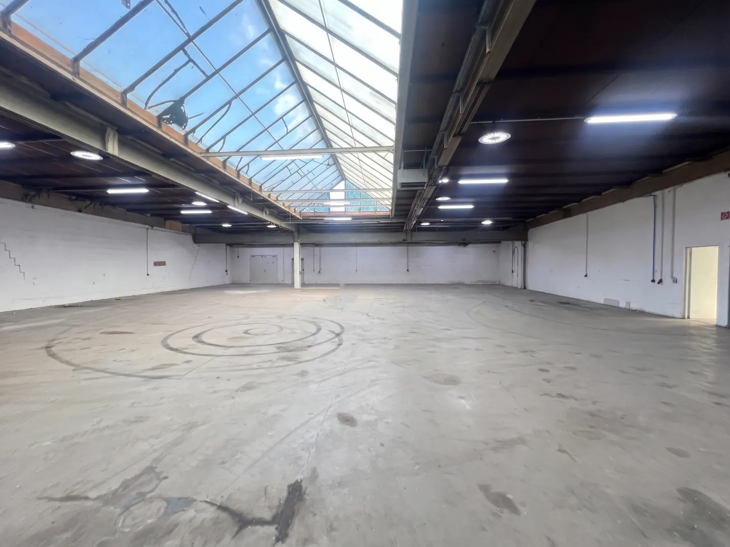 Empty warehouse space with white walls, concrete floor, and skylight. Tire marks on the floor.