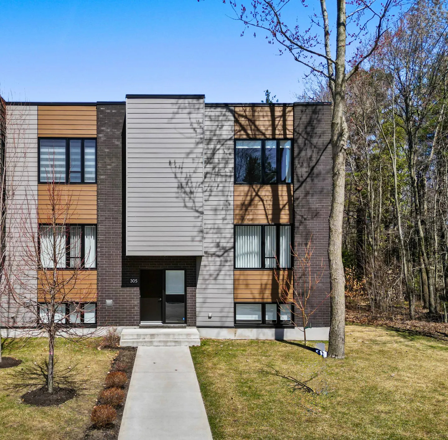 Modern two-story townhouse with gray, brown, and brick facade. A concrete walkway leads to the black front door. Trees in the background.