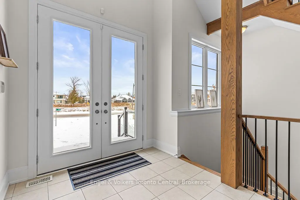 Bright foyer with white double doors leading to a snowy landscape. A striped mat lies inside. Stairs with wood and black railings are to the right.