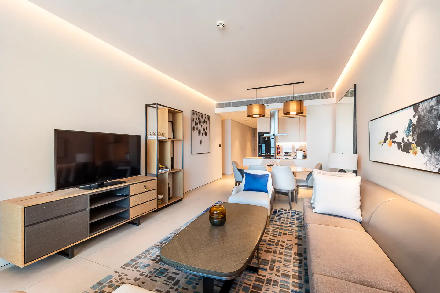 A modern living room with a TV, sofa, coffee table, and dining area. The color palette is neutral with blue accents.