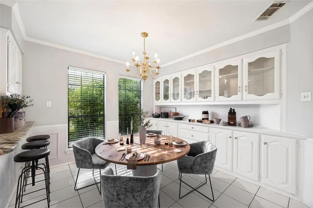 Bright kitchen with a round wood table, gray chairs, and white cabinets. A gold chandelier hangs above the table. Two windows with blinds let in natural light.