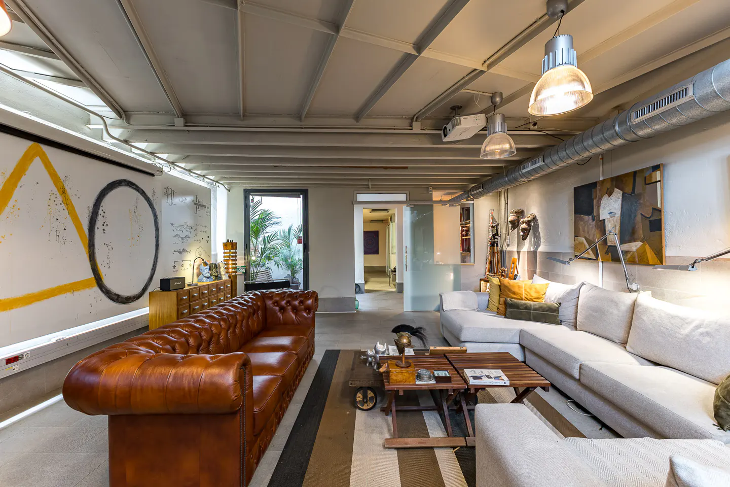 Modern industrial living room with a brown leather tufted sofa, white sectional, wooden coffee tables, and abstract art.