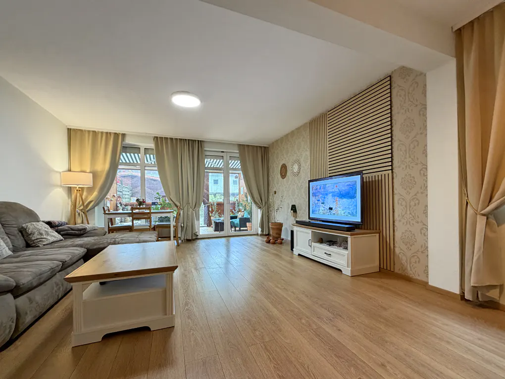 Living room with wood floors, gray sofa, and a light wood coffee table. A TV sits on a white console against a patterned wall. Curtains frame a patio door.