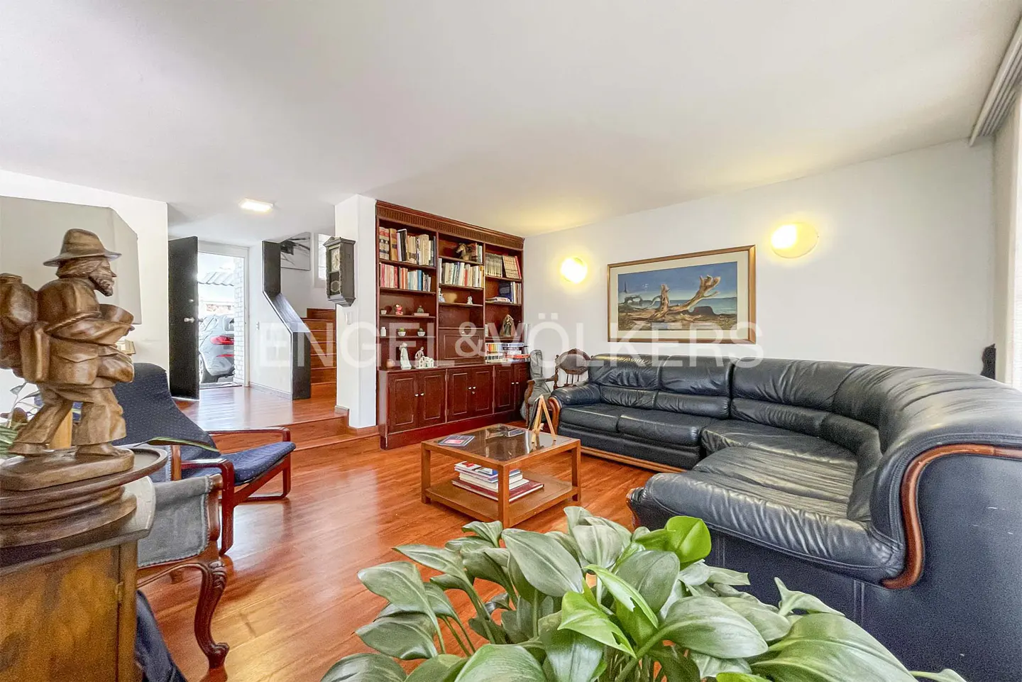 Living room with hardwood floors, a large black leather sectional, a wooden bookshelf, and a coffee table with books.