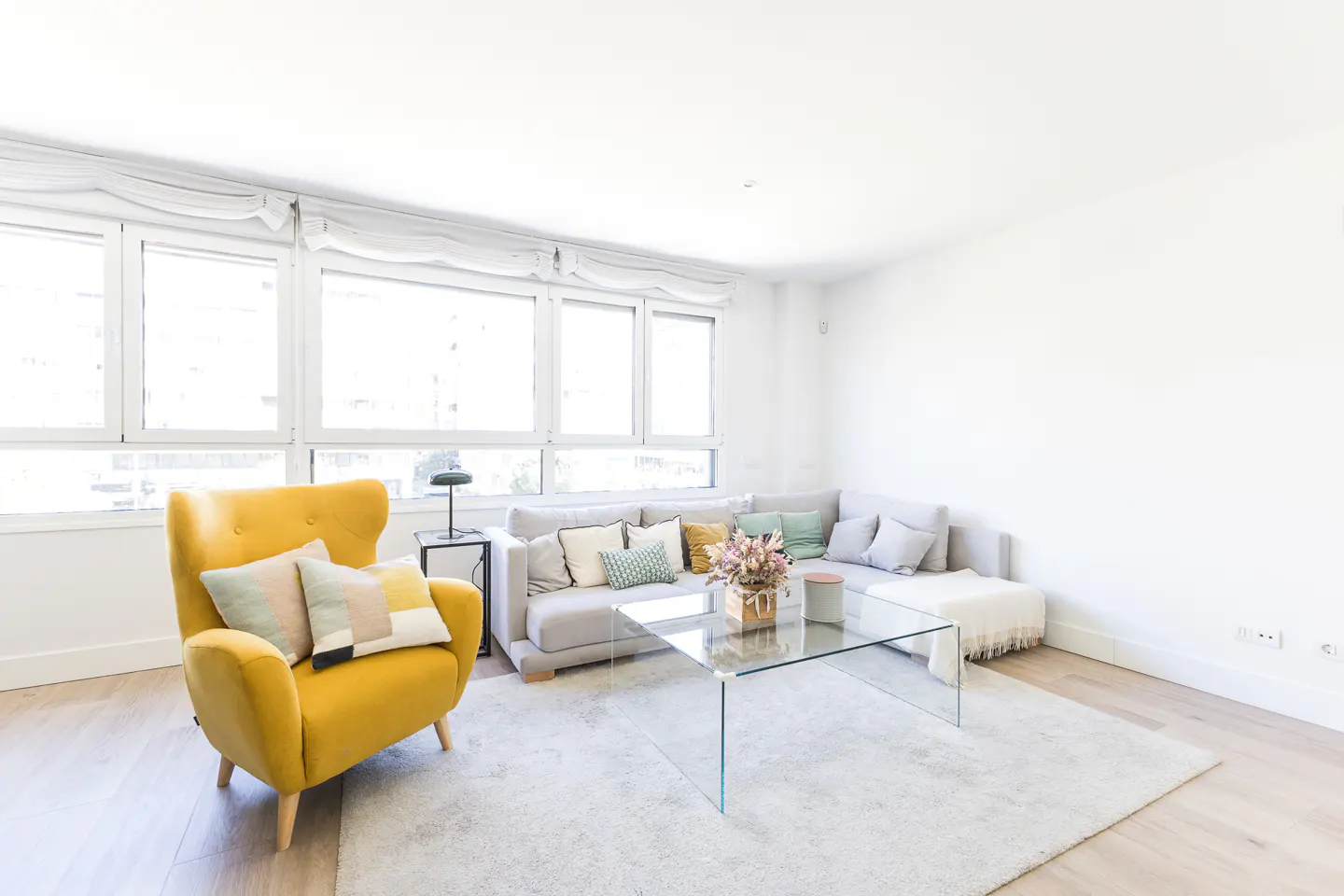 Bright living room with a yellow armchair, a gray sofa with pillows, and a glass coffee table on a white rug. Large windows provide natural light.