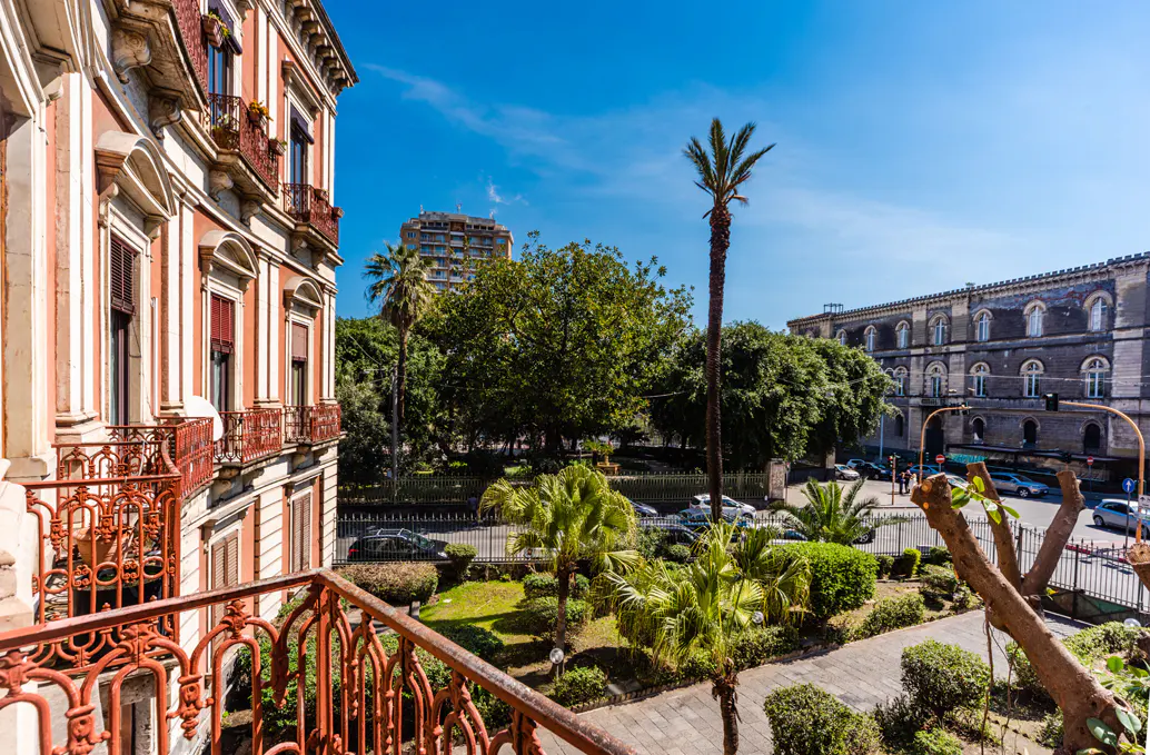 View from a balcony with red railings overlooking a park with trees and buildings under a blue sky.