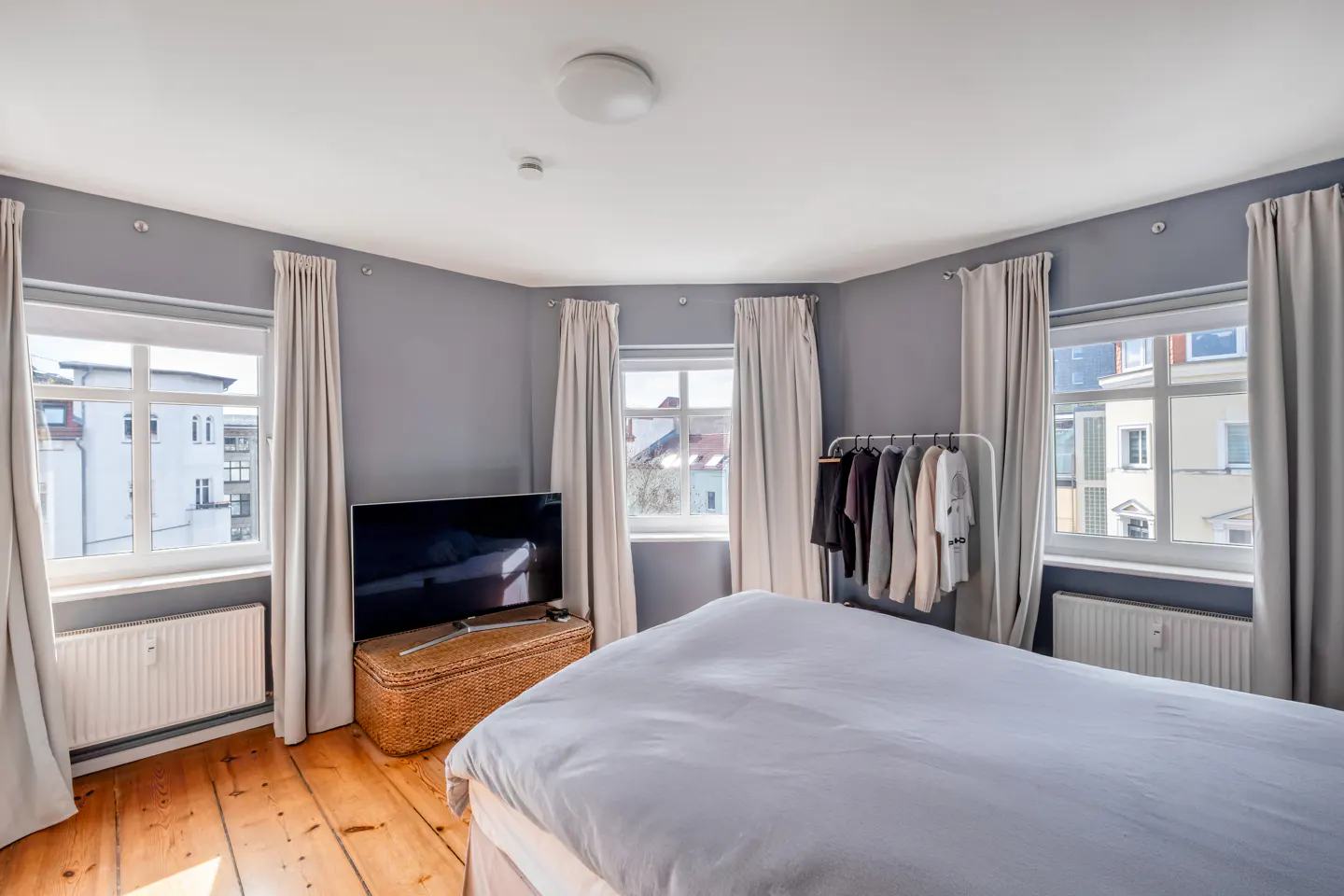 Bedroom with gray walls, wood floors, and white bedding. Windows with light curtains, a TV on a wicker stand, and a clothing rack are visible.