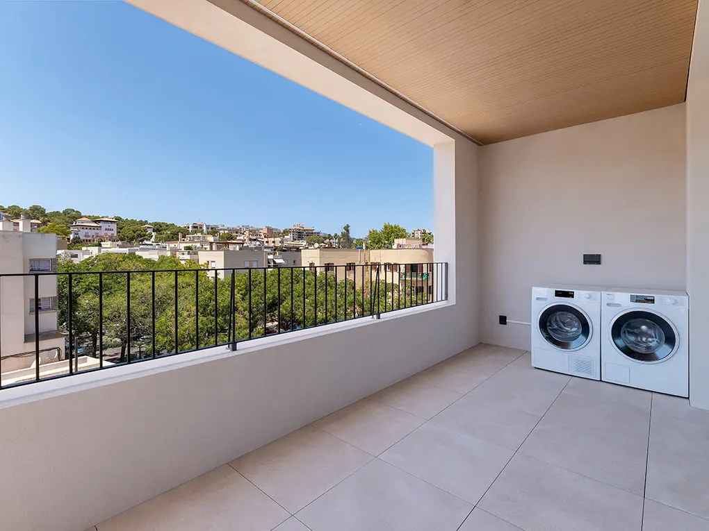 A bright balcony with a black railing overlooks a cityscape under a blue sky. Two white laundry machines sit against the wall.