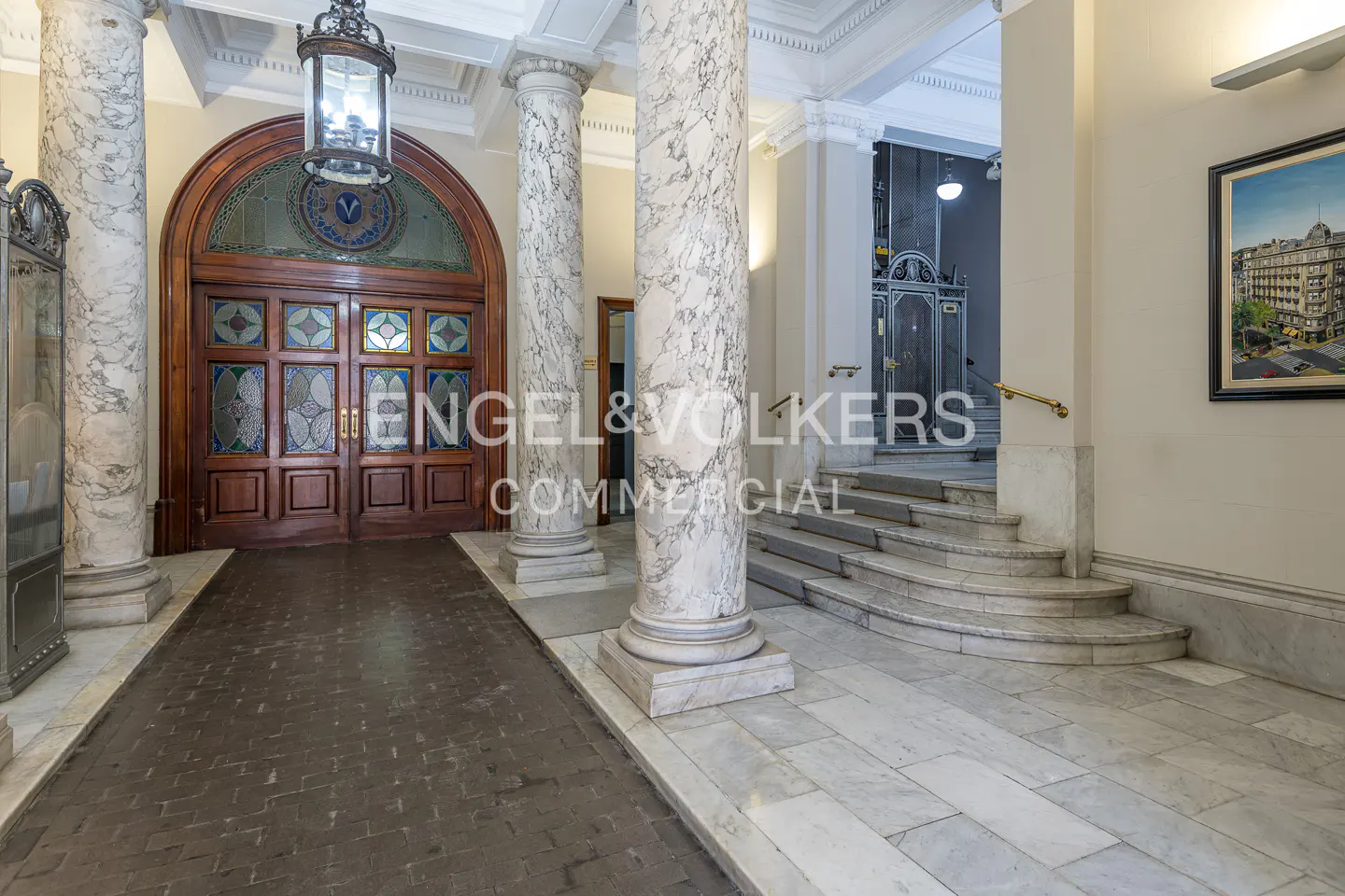 Elegant lobby with marble columns, arched wooden doors with stained glass, and a staircase. A vintage lantern hangs above.