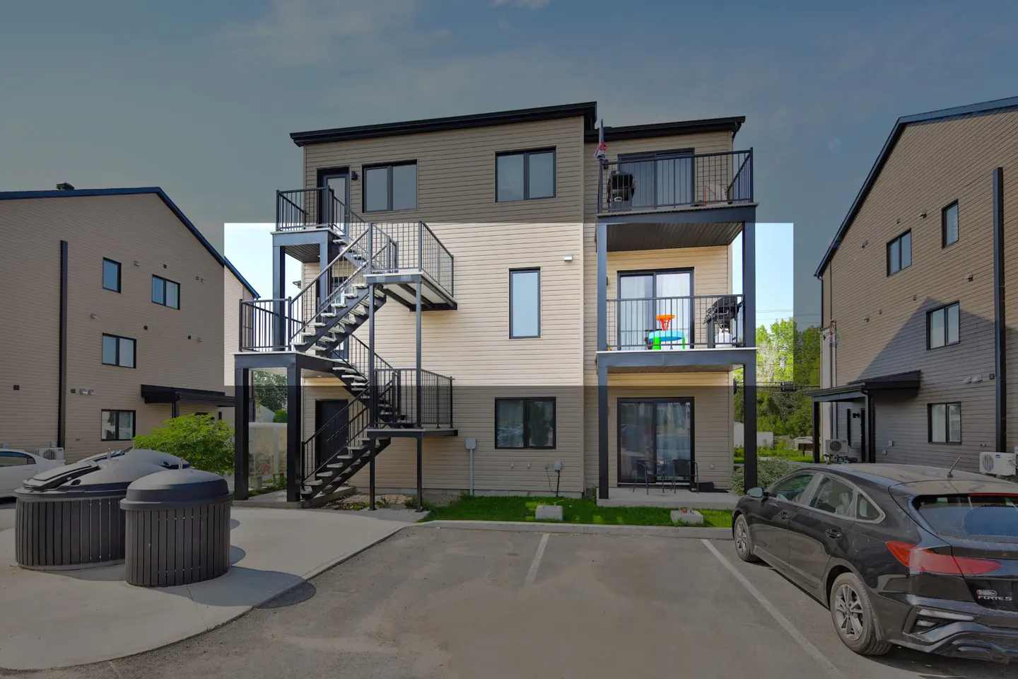 Tan apartment building with black stairs and balconies. A black car is parked in front.