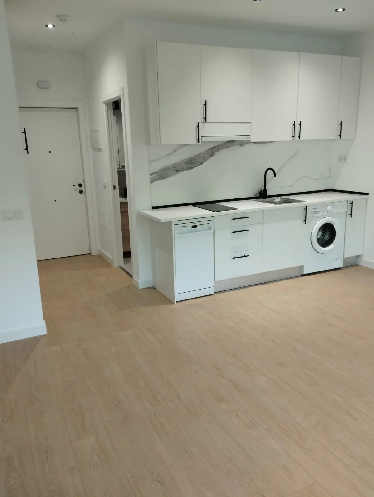 Bright, modern apartment interior with light wood floors, white walls, and a white kitchen with black hardware and marble backsplash.