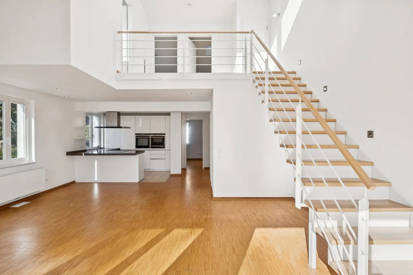 Bright, modern home interior with wood floors, white walls, and a staircase leading to a loft overlooking a kitchen.