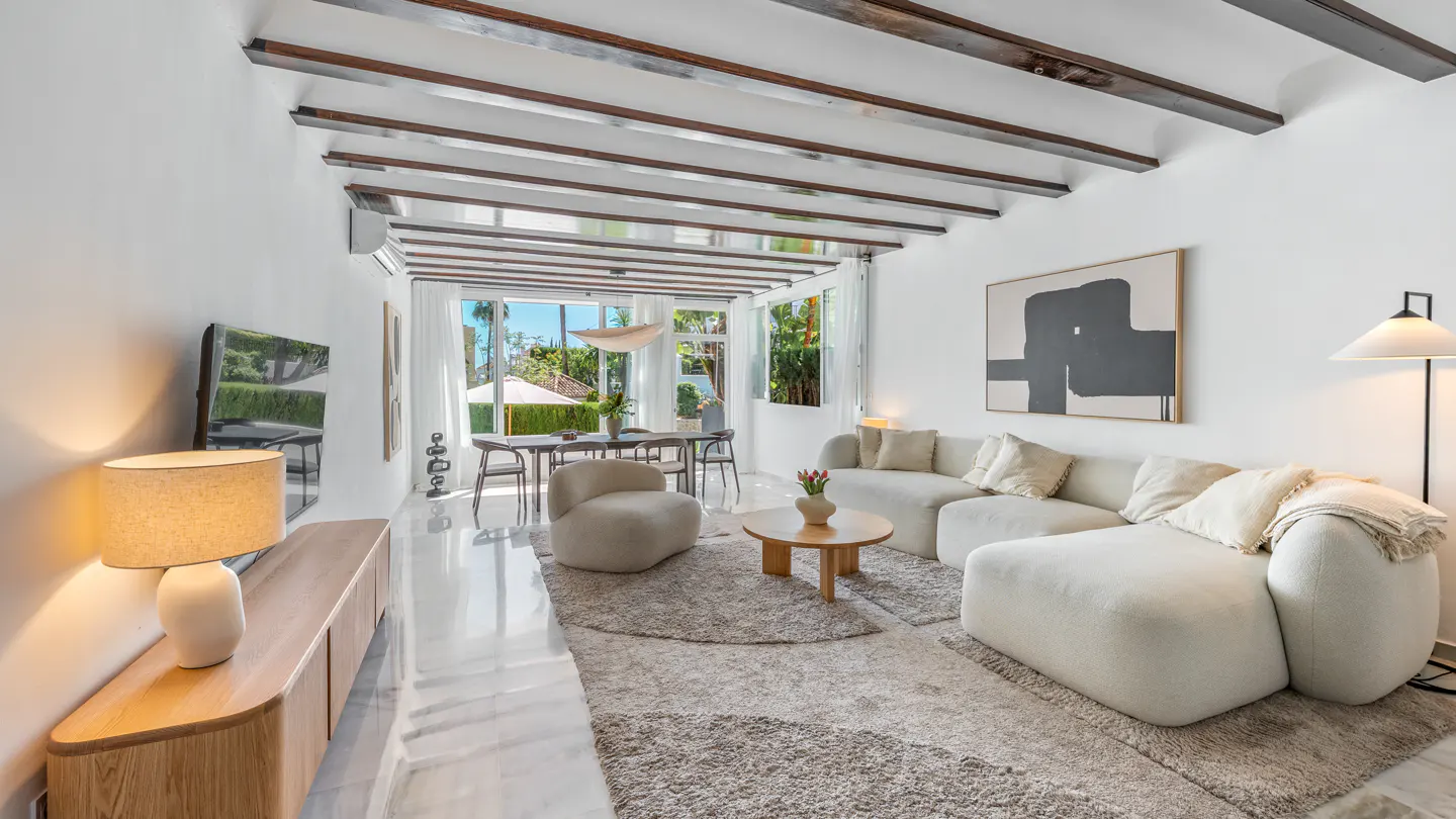 Bright living room with white walls, wood beams, and marble floors. A large white sectional sofa faces a round wood coffee table. Dining table visible through open doors.