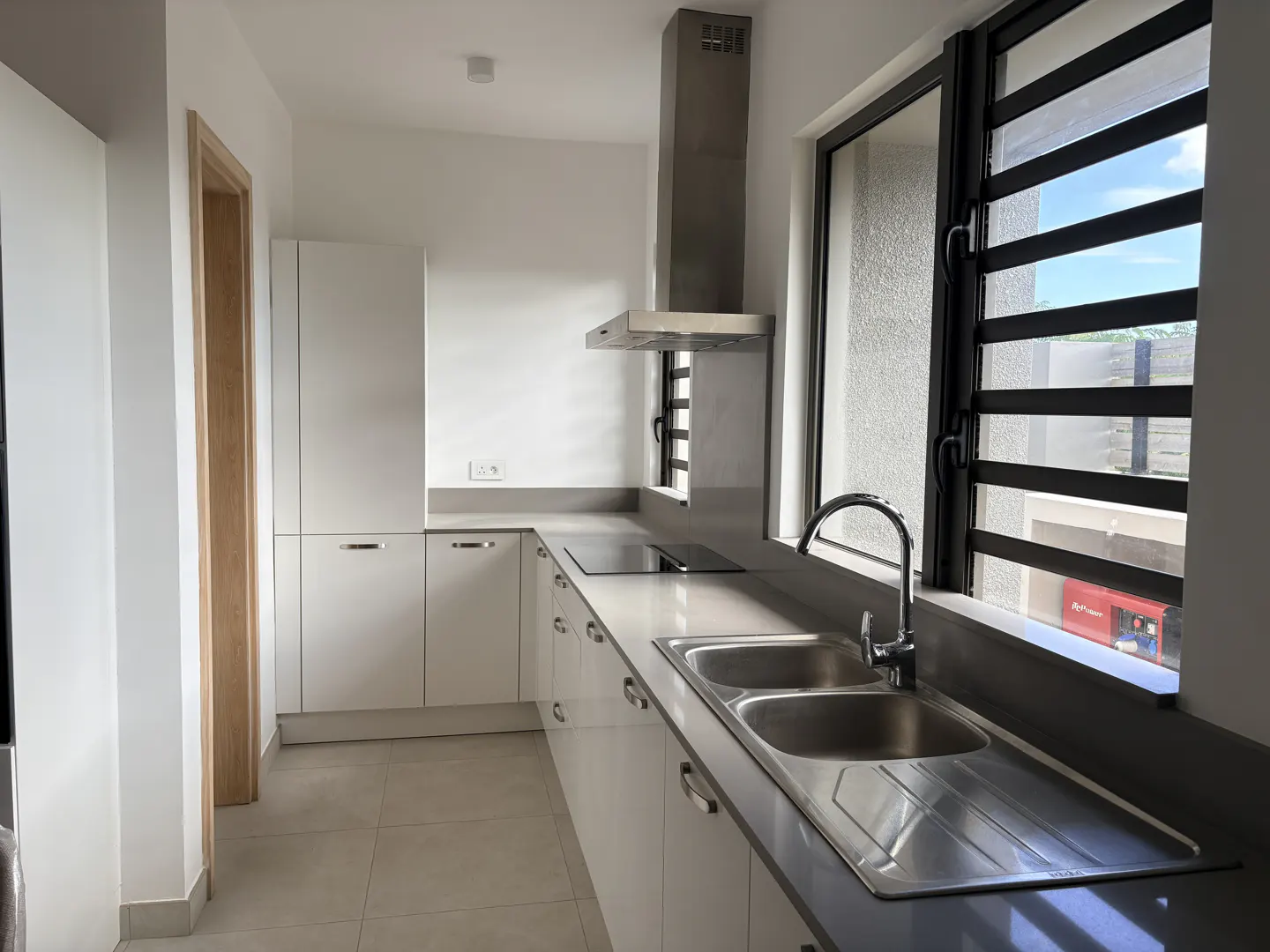 Bright kitchen with white cabinets, gray countertops, and stainless steel sink. A window with louvers offers a view of the outside.