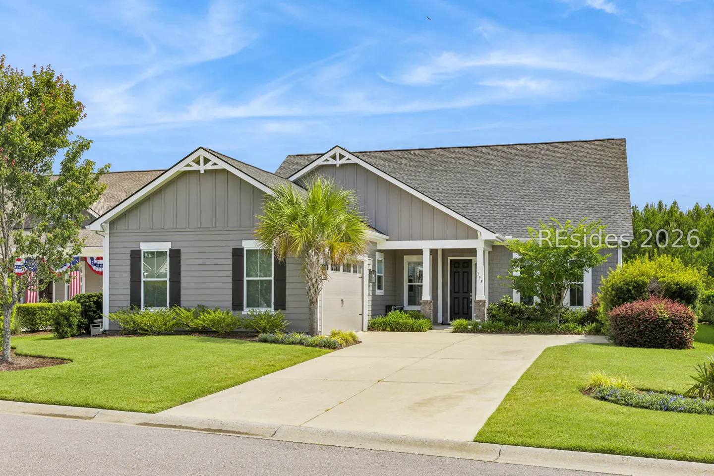 Single-story gray house with black shutters, a palm tree in the front yard, and a concrete driveway under a blue sky.