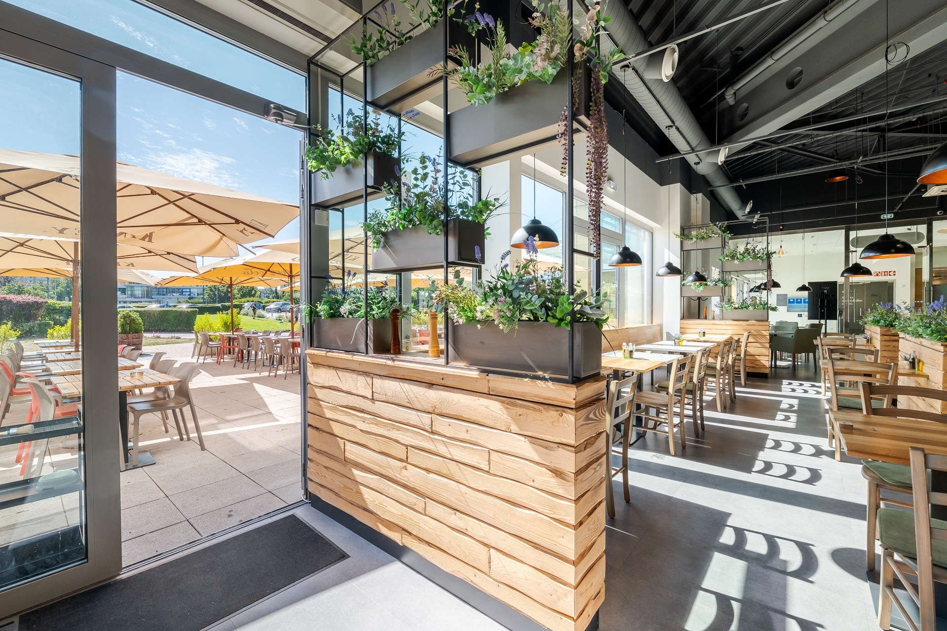 Modern restaurant interior with wooden partition, hanging plants, and an outdoor patio with umbrellas on a sunny day.