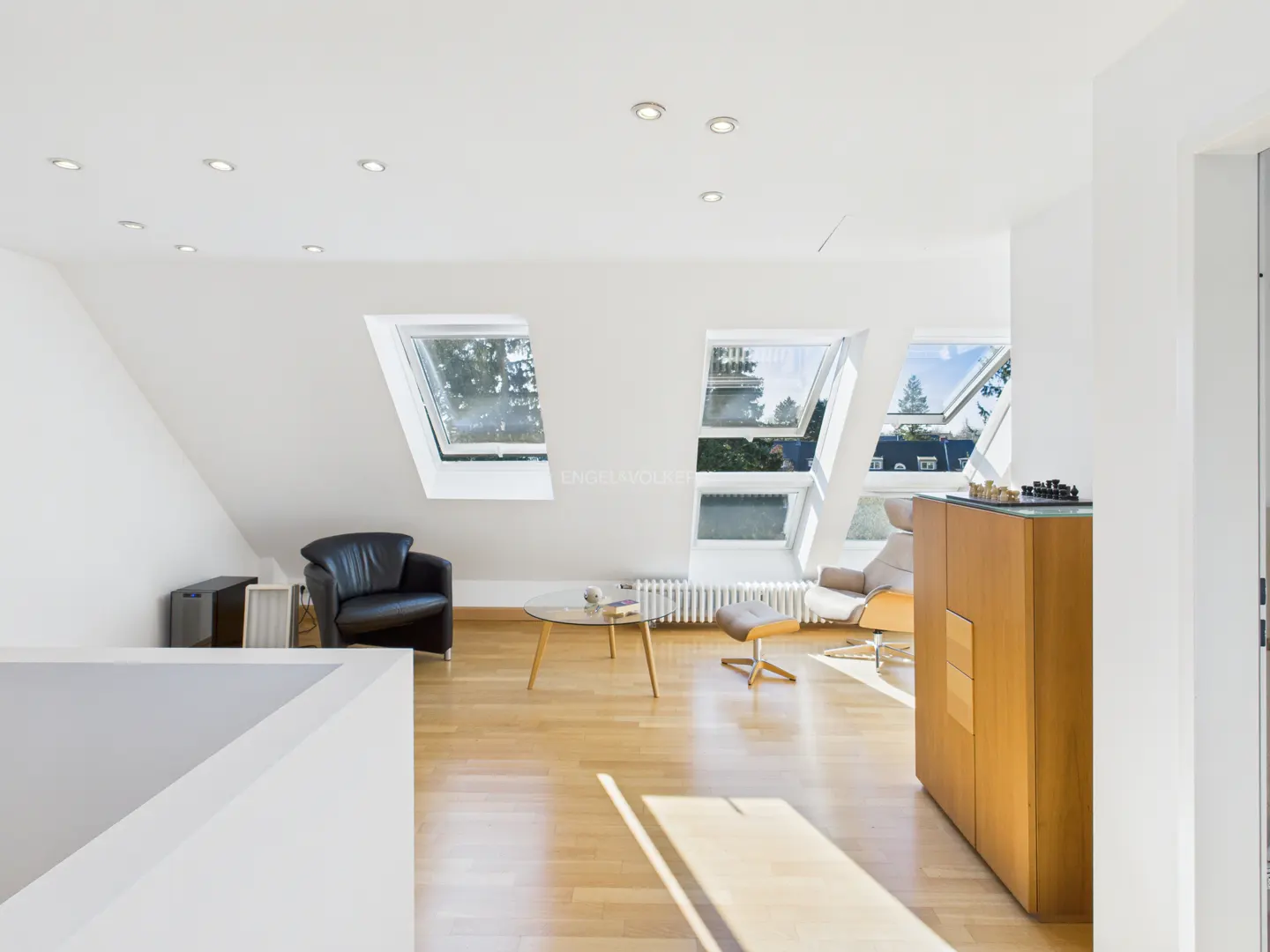 Bright attic living room with hardwood floors, white walls, and skylight windows. A black leather chair and wooden cabinet furnish the space.