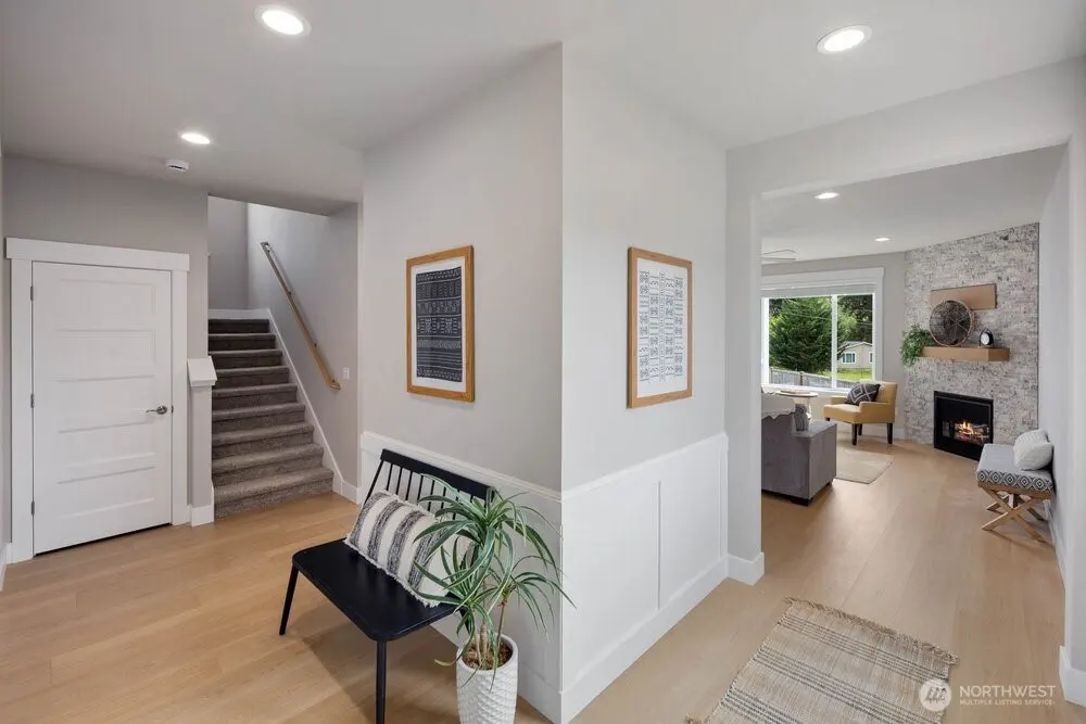Bright foyer with light wood floors, gray walls, and white trim. A staircase, bench, and living room with fireplace are visible.