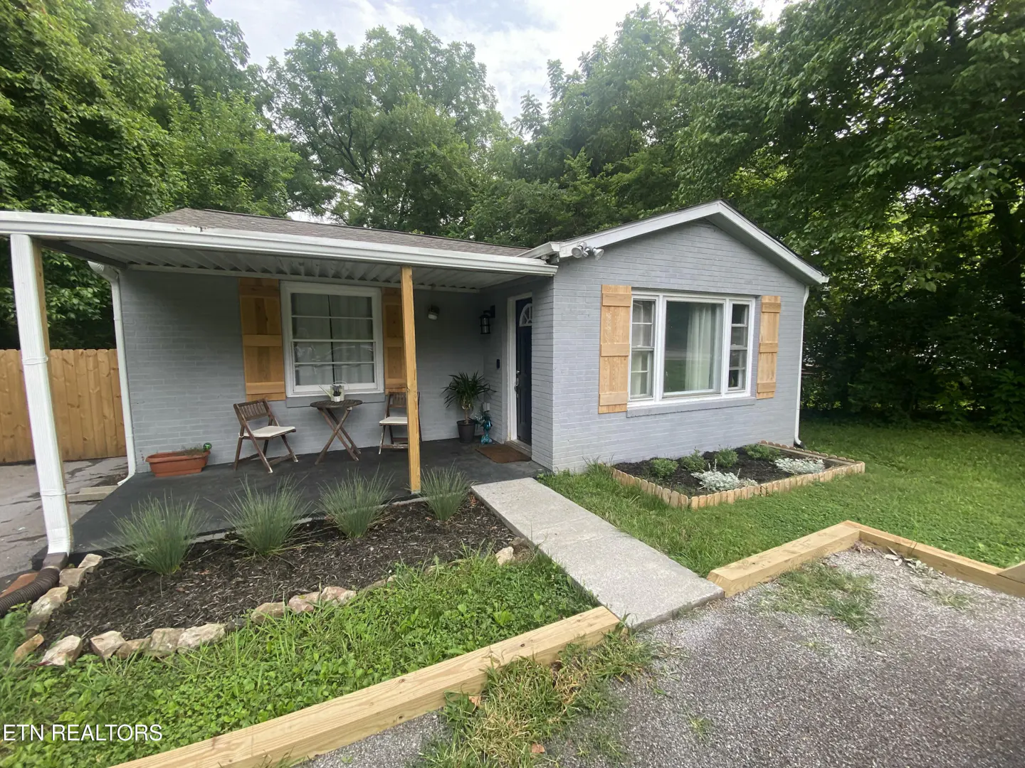 Exterior view of a gray brick house with light brown shutters and a small porch with chairs and a table.