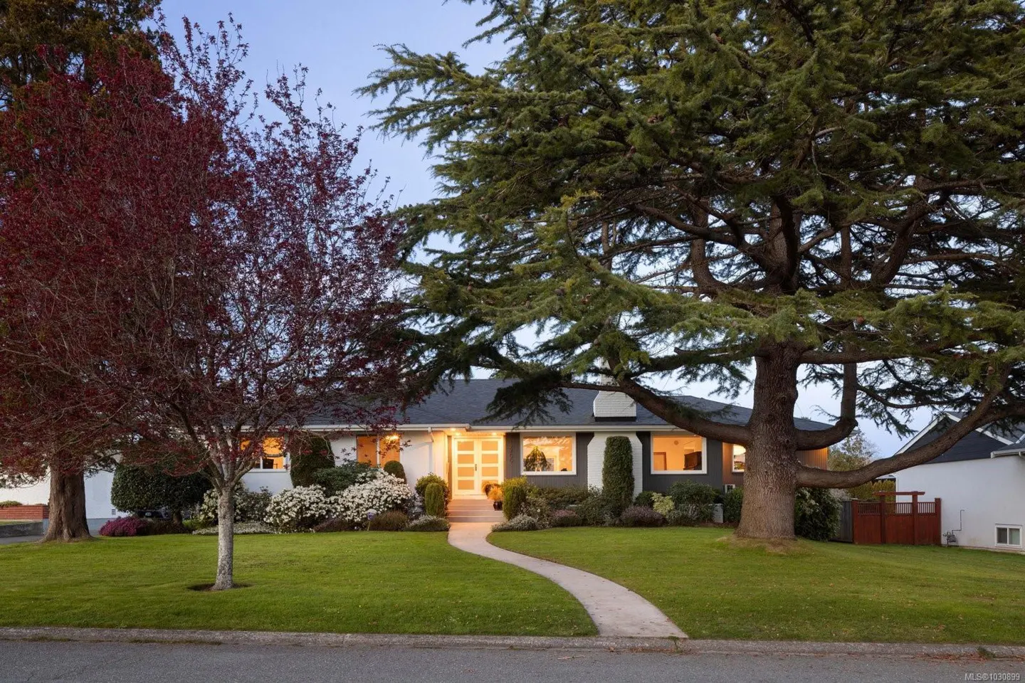 A single-story home with a dark roof, white trim, and a curved walkway leading to the front door. Two large trees frame the house.