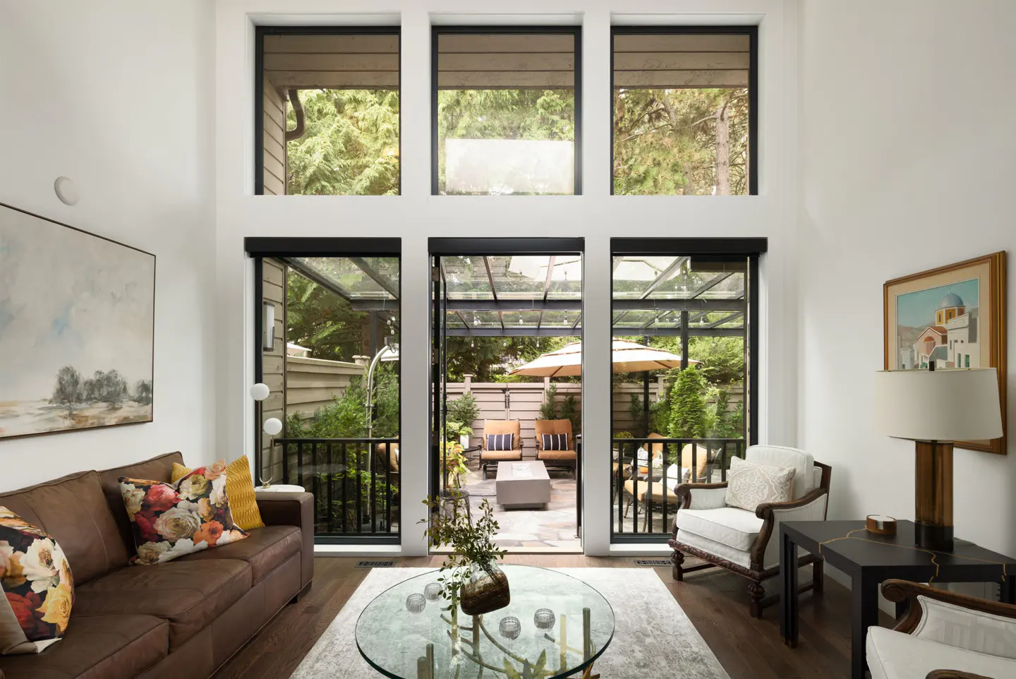 Bright living room with brown leather sofa, glass coffee table, and open doors to a patio with outdoor seating.