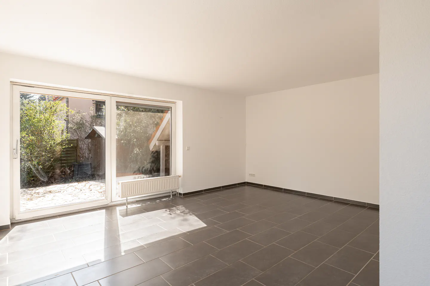 Bright, empty room with white walls and dark gray tiled floor. Large sliding glass doors lead to a garden with trees and a shed. A white radiator sits beneath the window.