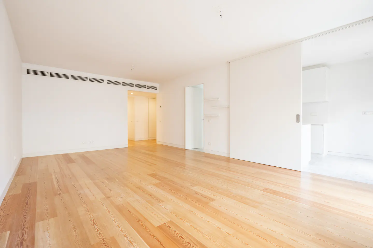 Bright, empty room with light wood floors and white walls. A sliding door leads to another room.