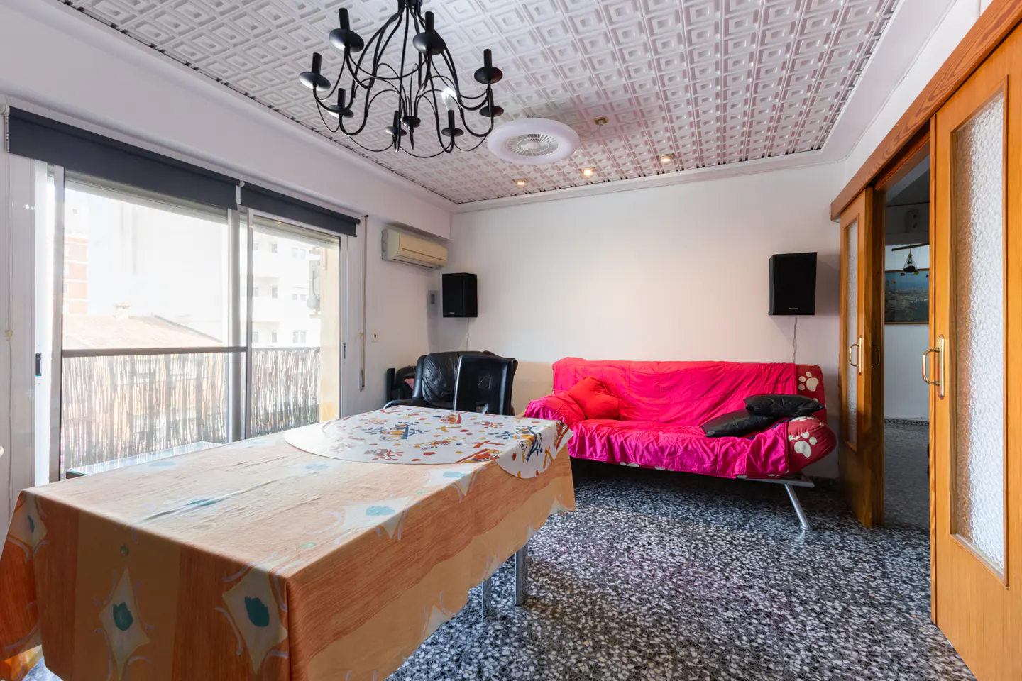 A living room with a table, pink sofa, black speakers, and a balcony. The ceiling has a decorative pattern and a black chandelier.