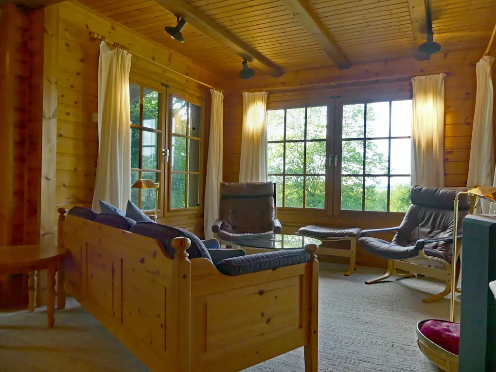 Cozy wood cabin interior with large windows, white curtains, and comfortable seating. Natural light floods the room.