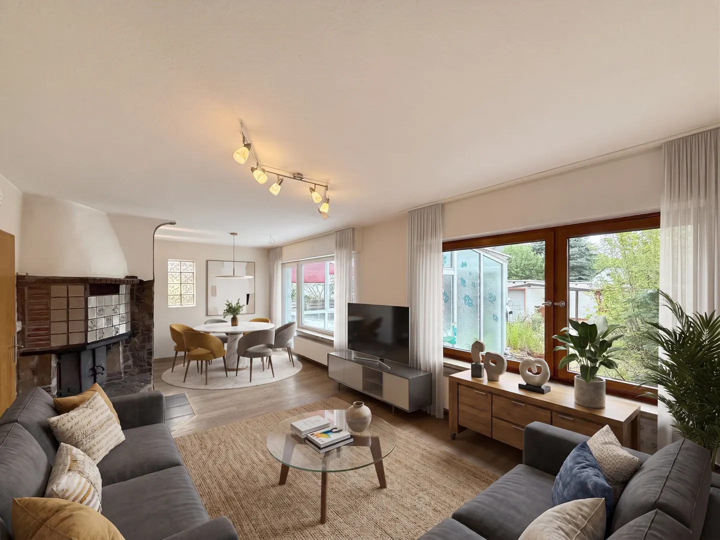 A bright living room with a gray sofa, fireplace, dining table, TV, and large windows.
