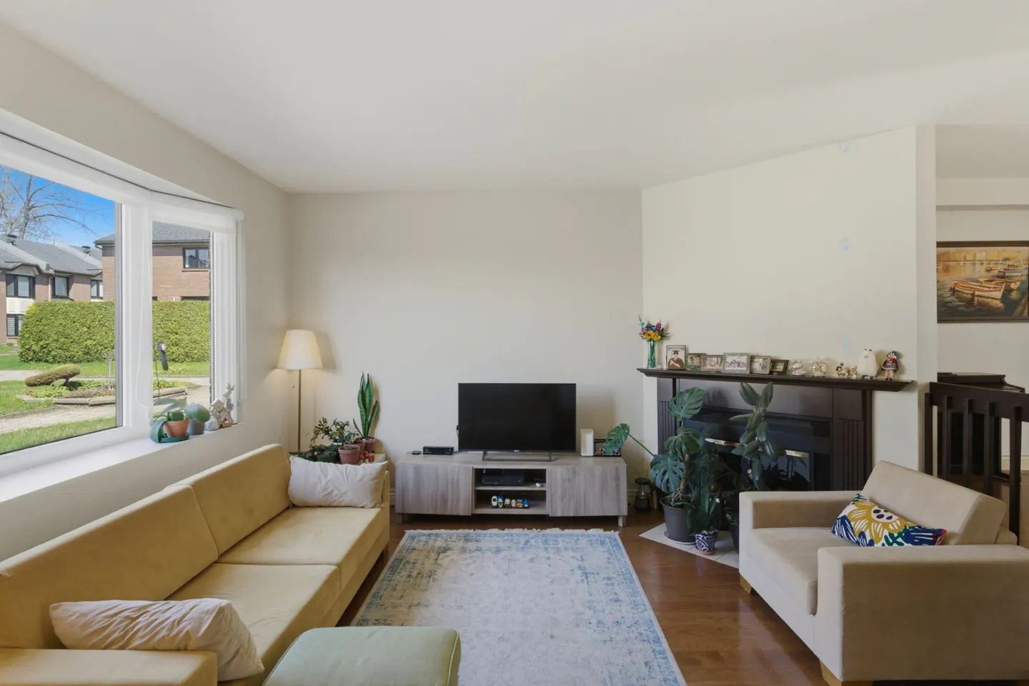 Living room with a yellow sofa, beige chair, TV, fireplace, and a large window with a view of a green lawn.