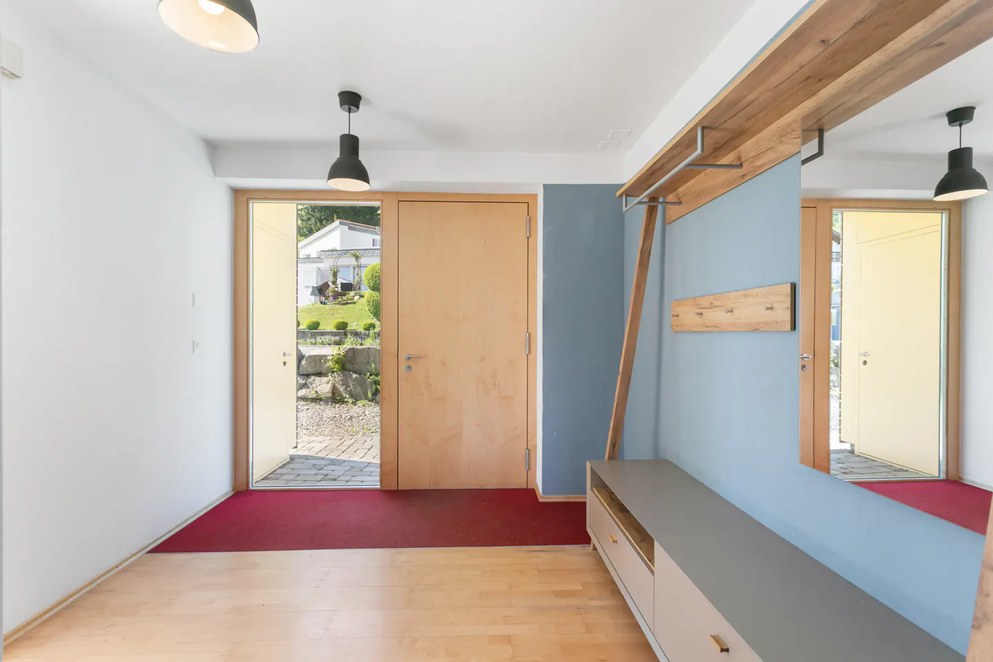 Bright foyer with light wood floors, white walls, and a red rug. An open door reveals a garden. A wooden coat rack and gray cabinet are on the right.