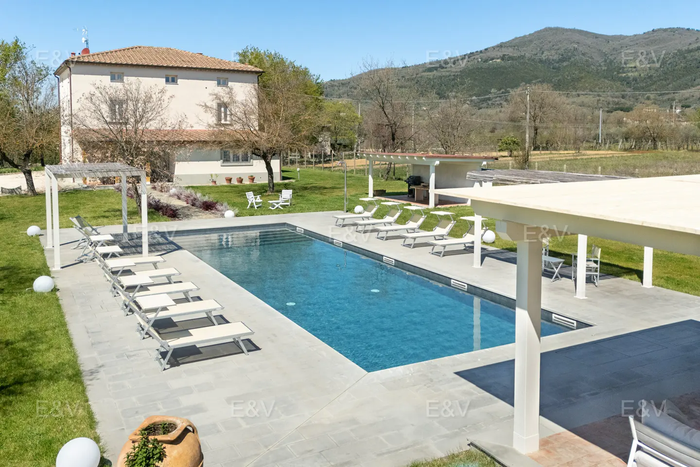 A rectangular blue pool is surrounded by white lounge chairs and a stone patio. A two-story house is in the background.