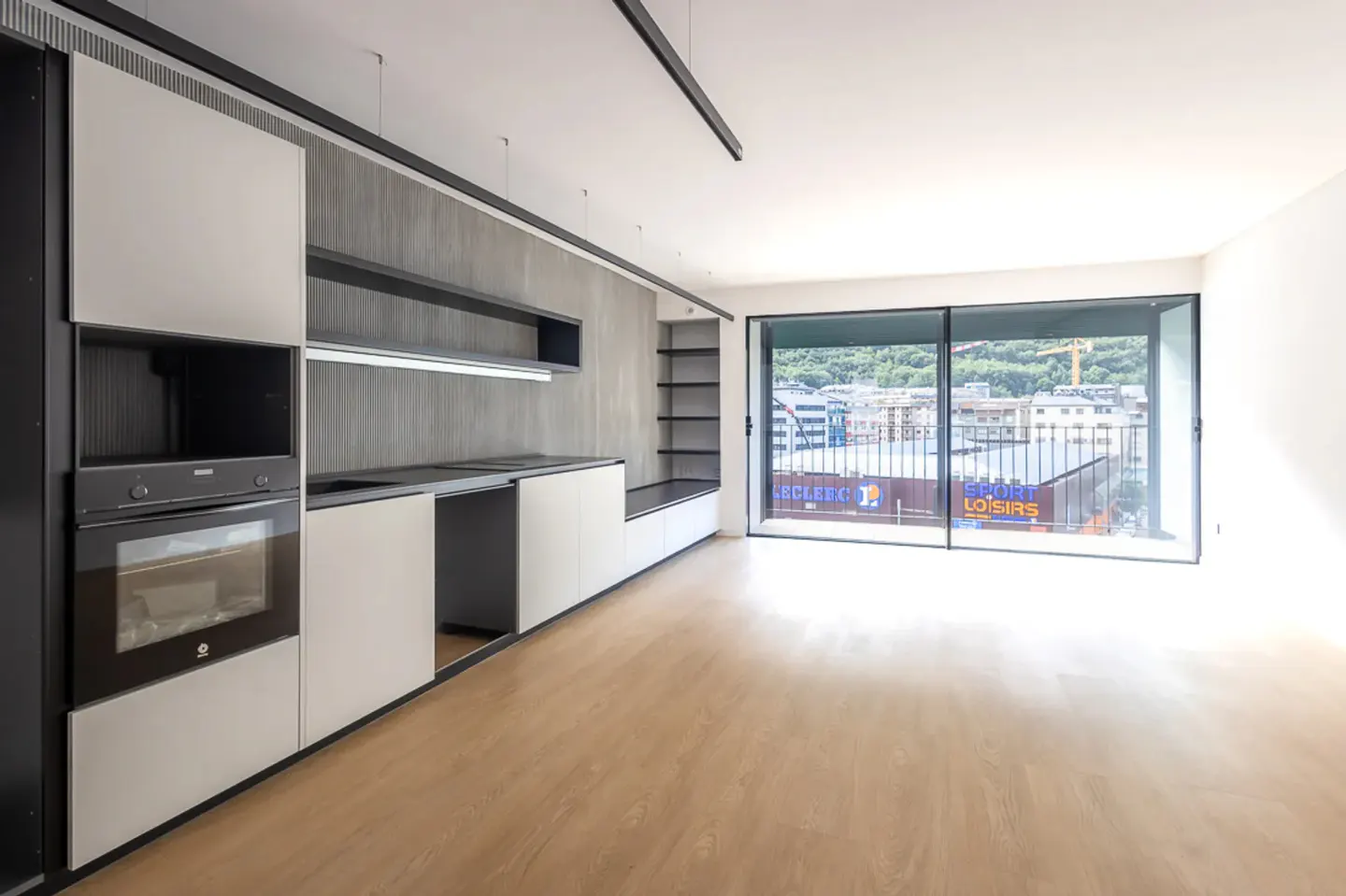 Modern kitchen with light wood floors, grey cabinetry, built-in oven, and large windows overlooking a city street.