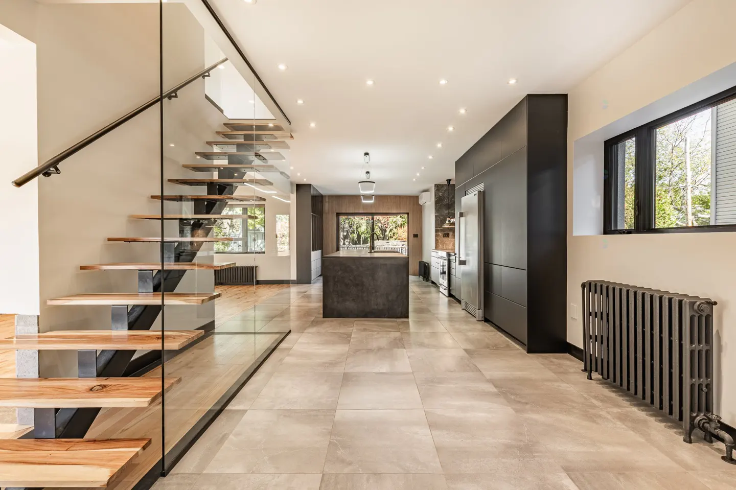 Open-concept home interior with floating wood stairs, glass railing, and a dark kitchen island on light tile flooring.