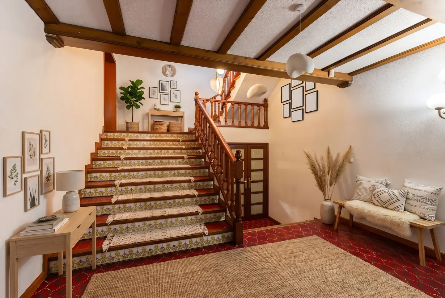 Interior view of a home foyer with a staircase, wooden beams, and neutral decor. A bench with pillows sits near a vase of pampas grass.