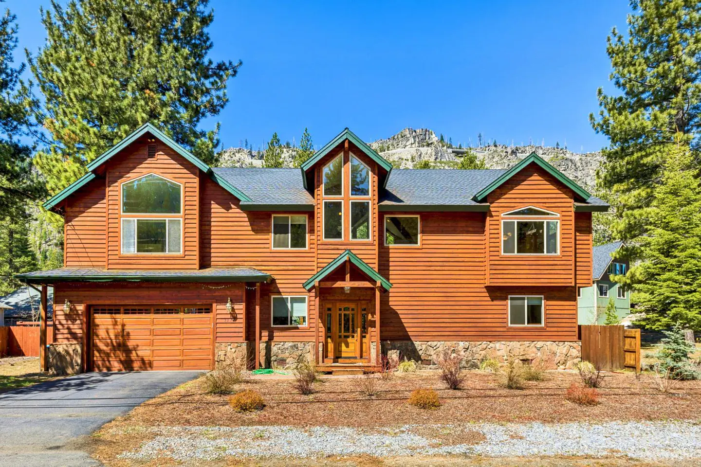 Two-story wood house with green trim, a gray roof, and a two-car garage, surrounded by trees and a blue sky.