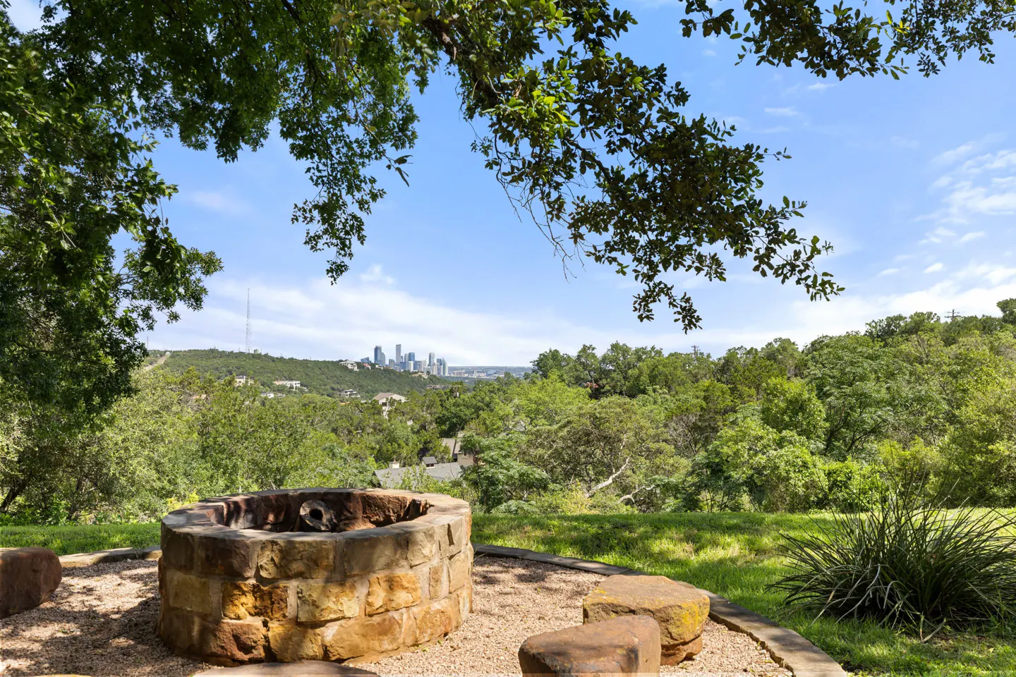 Stone fire pit in a grassy backyard overlooking a lush green hillside with a distant city skyline under a blue sky.