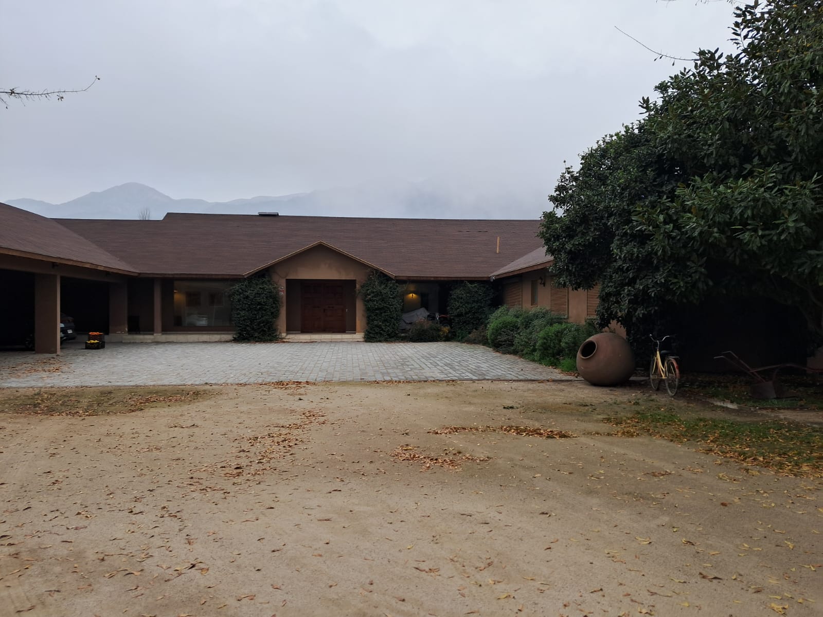 Single-story house with a brown roof, paved driveway, large clay pot, and bicycle, set against misty mountains.
