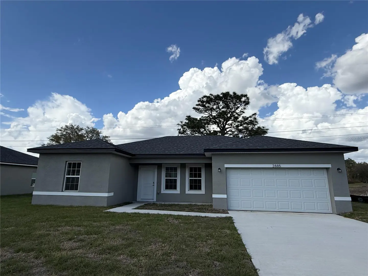 A single-story gray house with a white garage door and a dark roof under a blue sky with white clouds.