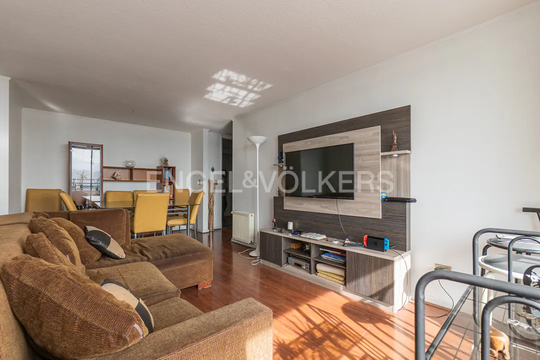 Living room with brown sofa, yellow chairs, and a TV mounted on a gray and brown wall unit. Hardwood floors and white walls.
